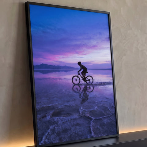 Ciclista em silhueta pedalando na água rasa sob céu roxo e rosa do pôr do sol, com reflexo, quadro decorativo