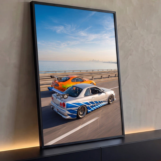 Carro prata com faixas azuis e carro laranja correndo em ponte à beira-mar sob céu azul, quadro decorativo