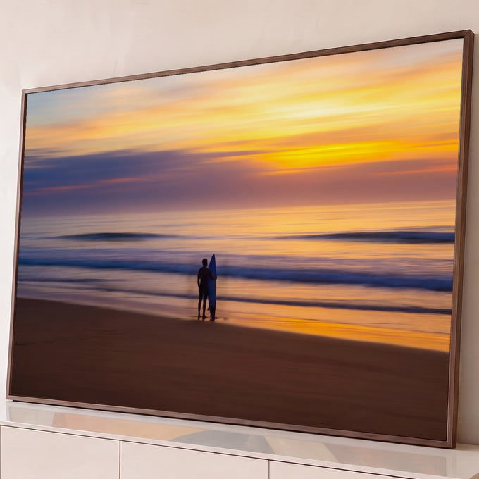 Silhueta de surfista com prancha azul na areia diante do mar ao pôr do sol laranja, quadro decorativo