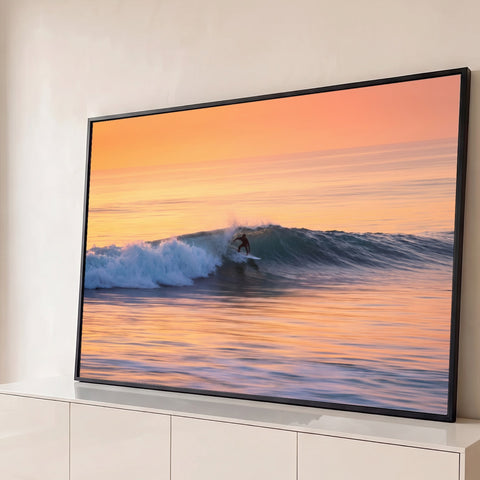 Surfista deslizando numa onda azul no mar ao pôr do sol, com céu laranja e reflexos dourados, quadro decorativo