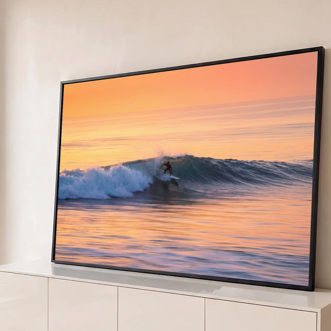 Surfista deslizando numa onda azul no mar ao pôr do sol, com céu laranja e reflexos dourados, quadro decorativo