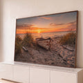 Barco de madeira rústico na areia entre capim de dunas com pôr do sol laranja no mar, quadro decorativo