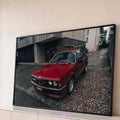 BMW coupé clássico vermelho estacionado em rua molhada com prédios cinza ao fundo, quadro decorativo
