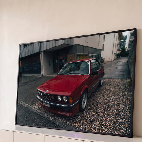 BMW coupé clássico vermelho estacionado em rua molhada com prédios cinza ao fundo, quadro decorativo