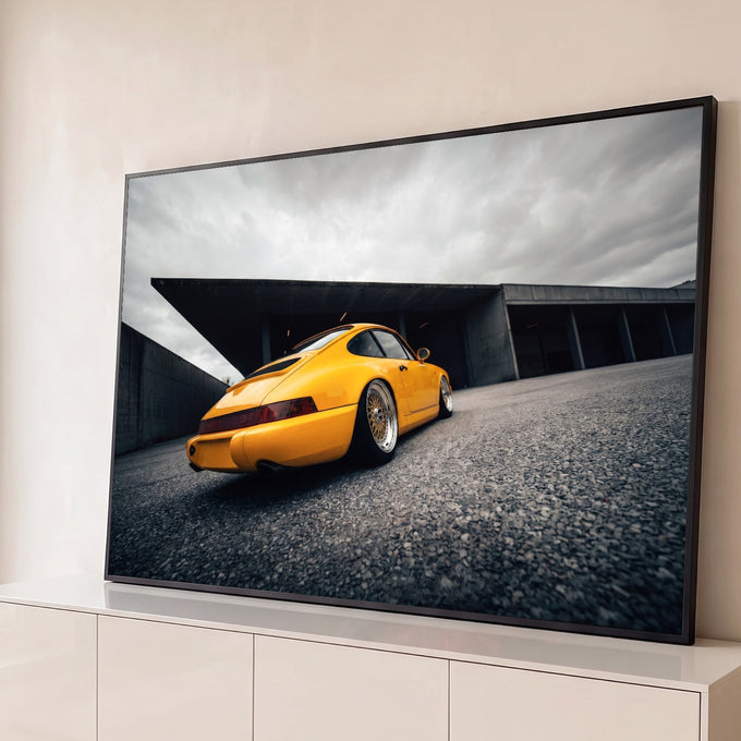 Porsche 911 amarelo visto de trás com rodas douradas sob céu nublado e concreto, quadro decorativo