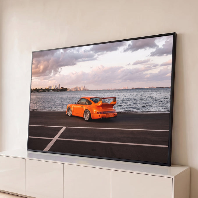 Porsche 911 laranja com aerofólio à beira-mar, skyline e céu nublado ao entardecer, quadro decorativo