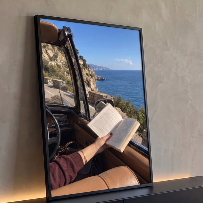 Mão segurando livro aberto em carro conversível retrô, estrada à beira-mar azul, quadro decorativo