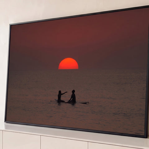 Sol vermelho se pondo no mar com casal de surfistas em silhueta sobre pranchas, quadro decorativo
