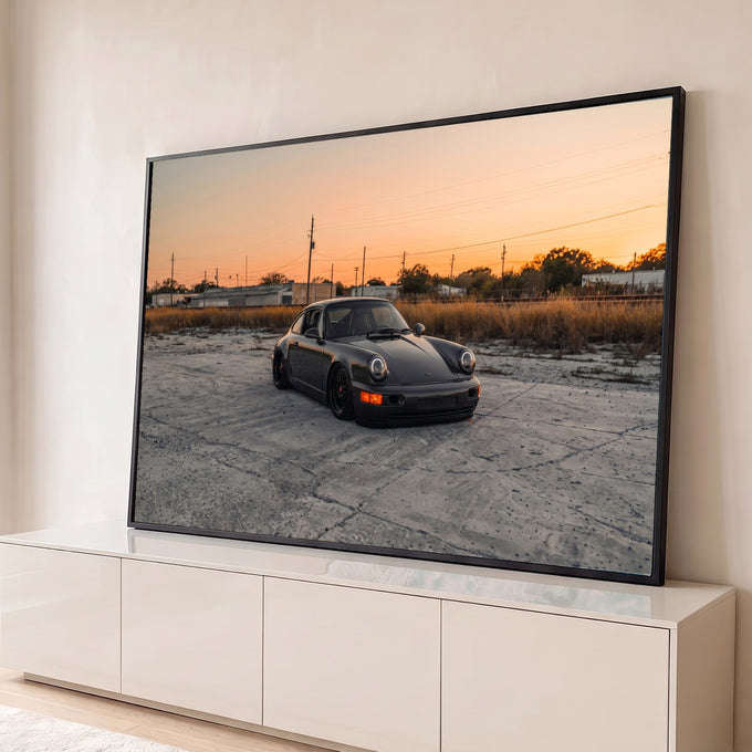 Porsche 911 preto rebaixado sobre concreto com céu alaranjado de pôr do sol, quadro decorativo