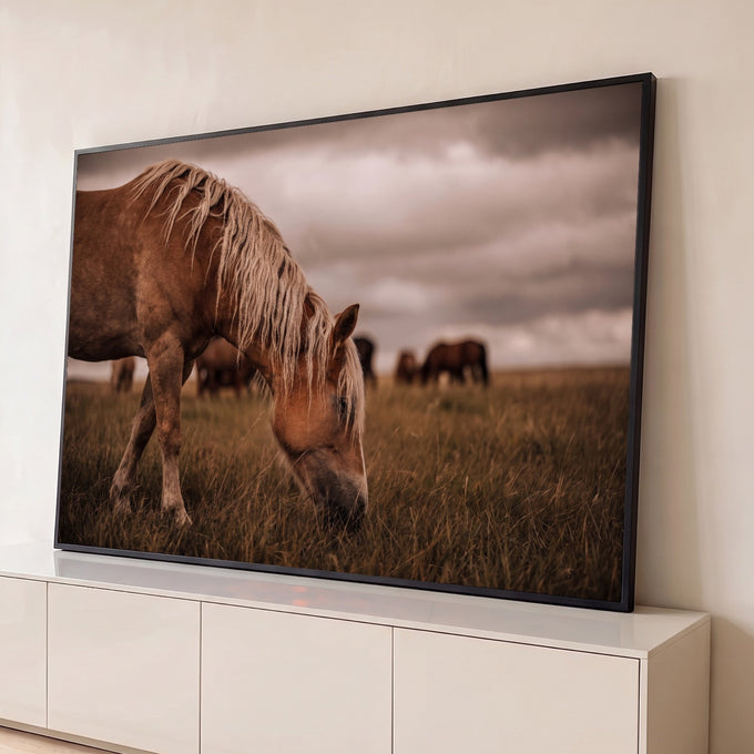 Cavalo de crina loira pastando em campo com manada ao fundo e céu nublado, quadro decorativo
