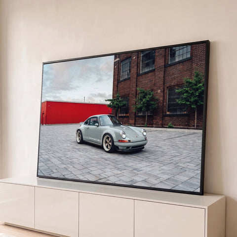 Porsche 911 clássico cinza com rodas douradas em pátio de pedras e prédio de tijolos, quadro decorativo