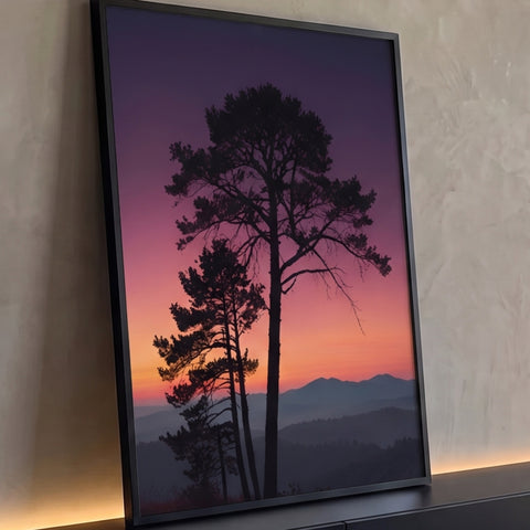 Pinheiros em silhueta contra céu roxo e laranja do pôr do sol sobre montanhas enevoadas, quadro decorativo