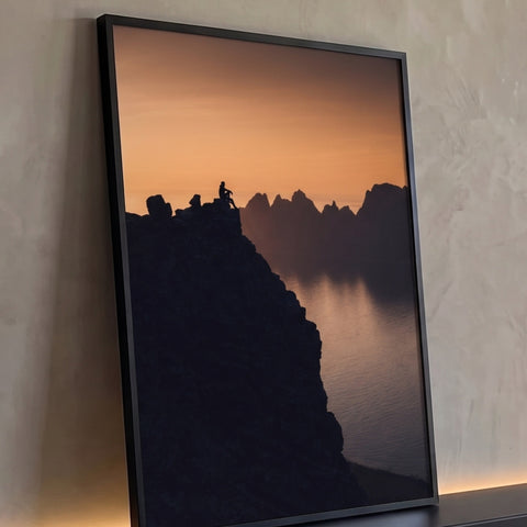 Silhueta de pessoa sentada em penhasco diante de montanhas e fiorde ao pôr do sol laranja, quadro decorativo