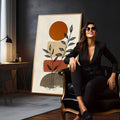Ramos de folhas em traço preto sobre sol laranja e formas terrosas em fundo bege, quadro decorativo