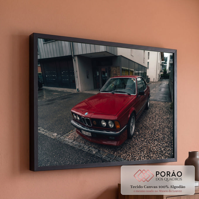 BMW coupé clássico vermelho estacionado em rua molhada com prédios cinza ao fundo, quadro decorativo