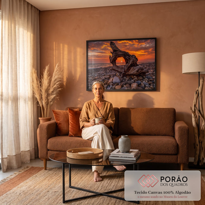 Tronco de madeira emoldurando o pôr do sol laranja sobre o mar em praia de seixos, quadro decorativo